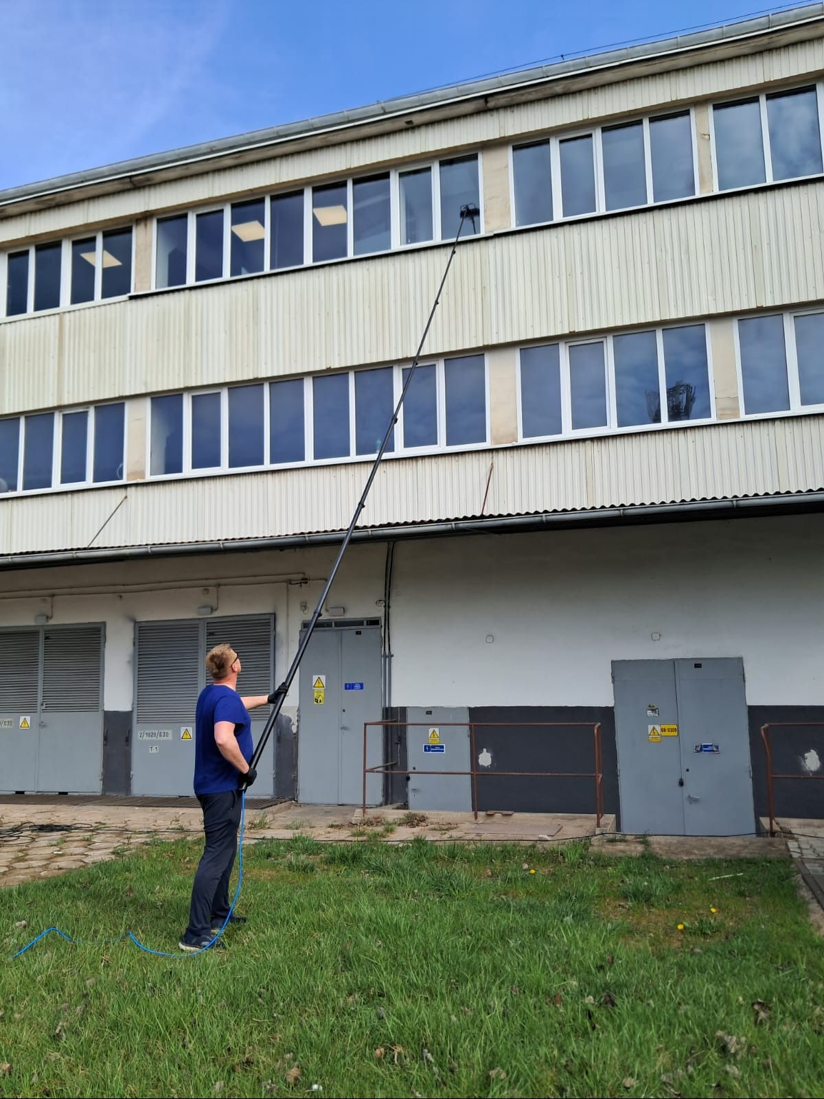 Mężczyzna myje okna na wysokości za pomocą długiej lancy. Budynek z szarymi drzwiami i zielonym trawnikiem w tle. Błękitne niebo.