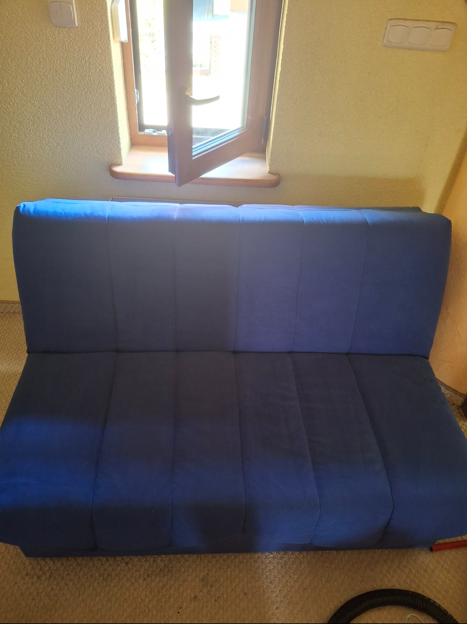Niebieska, rozłożona kanapa z widocznym, otwartym oknem w tle. Na podłodze widoczny fragment odkurzacza. Teksturowana ściana w tle.