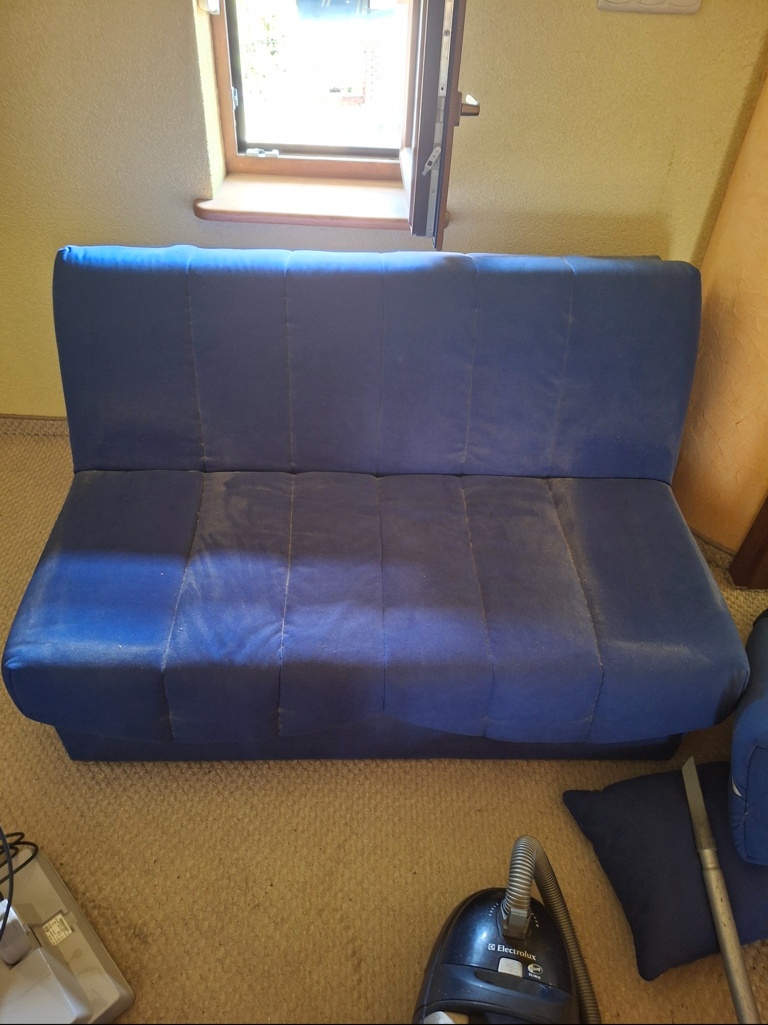 Niebieska, zabrudzona kanapa przed praniem. Widoczny odkurzacz Electrolux i poduszka. Ujęcie z góry, w tle okno.