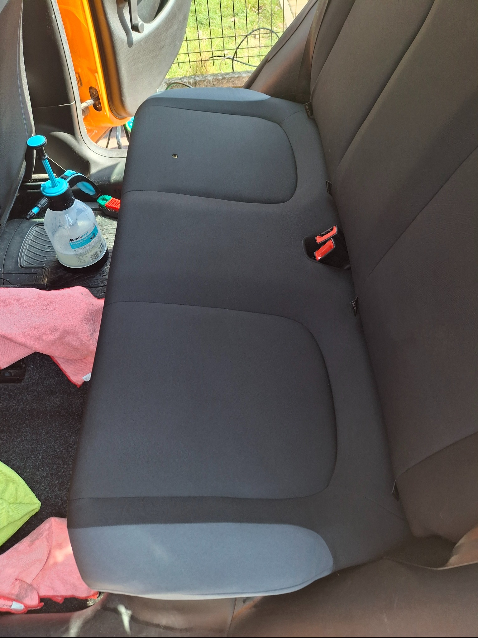 Wnętrze samochodu z widoczną tylną kanapą w trakcie czyszczenia, z użyciem spryskiwacza i ściereczek. Otwarta pomarańczowa klapka drzwi.