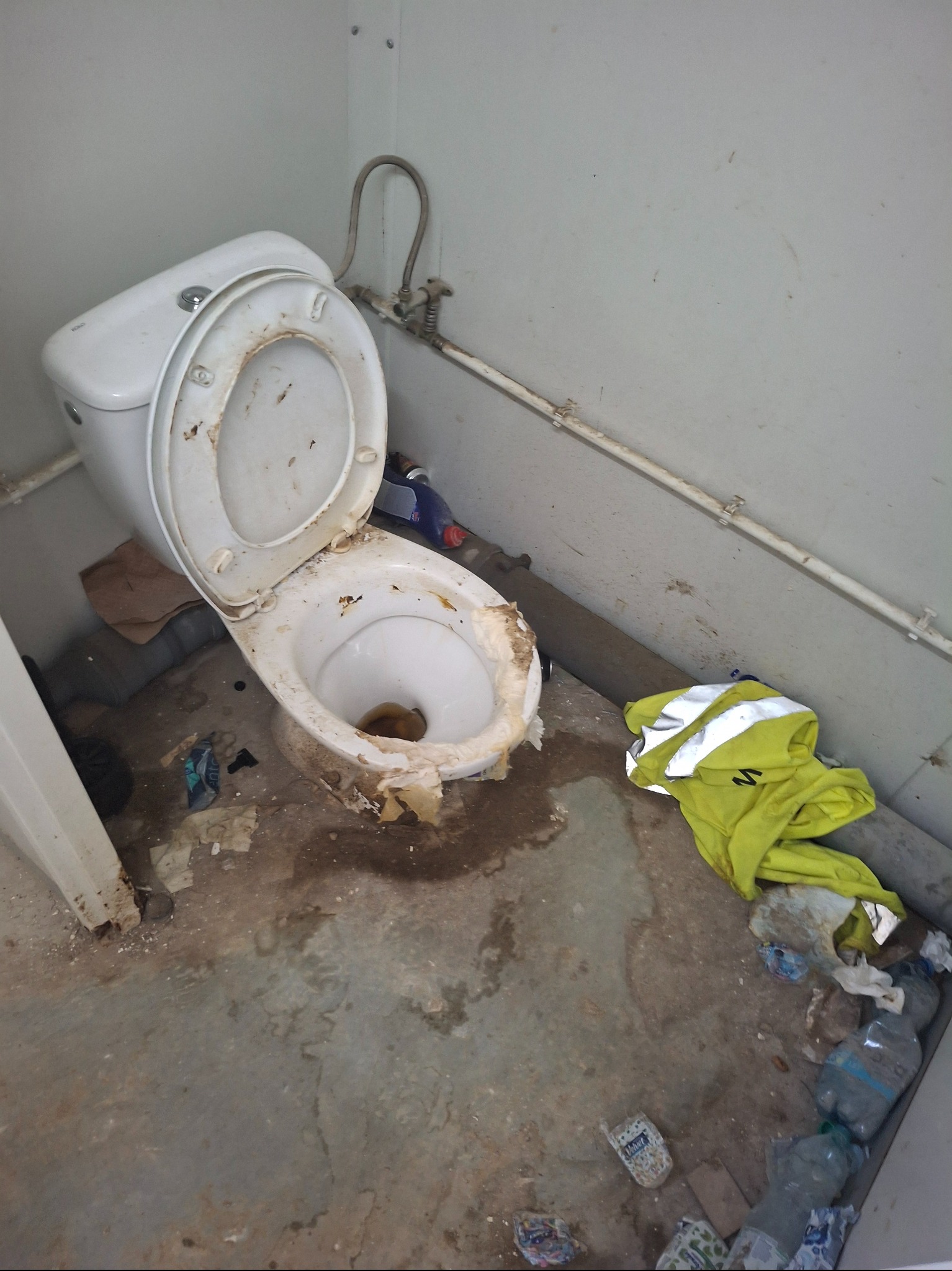 Bardzo zaniedbana toaleta z otwartą klapą, widoczne ślady brudu, resztki i śmieci na podłodze, obok leży żółta bluza robocza. Stan przed sprzątaniem.