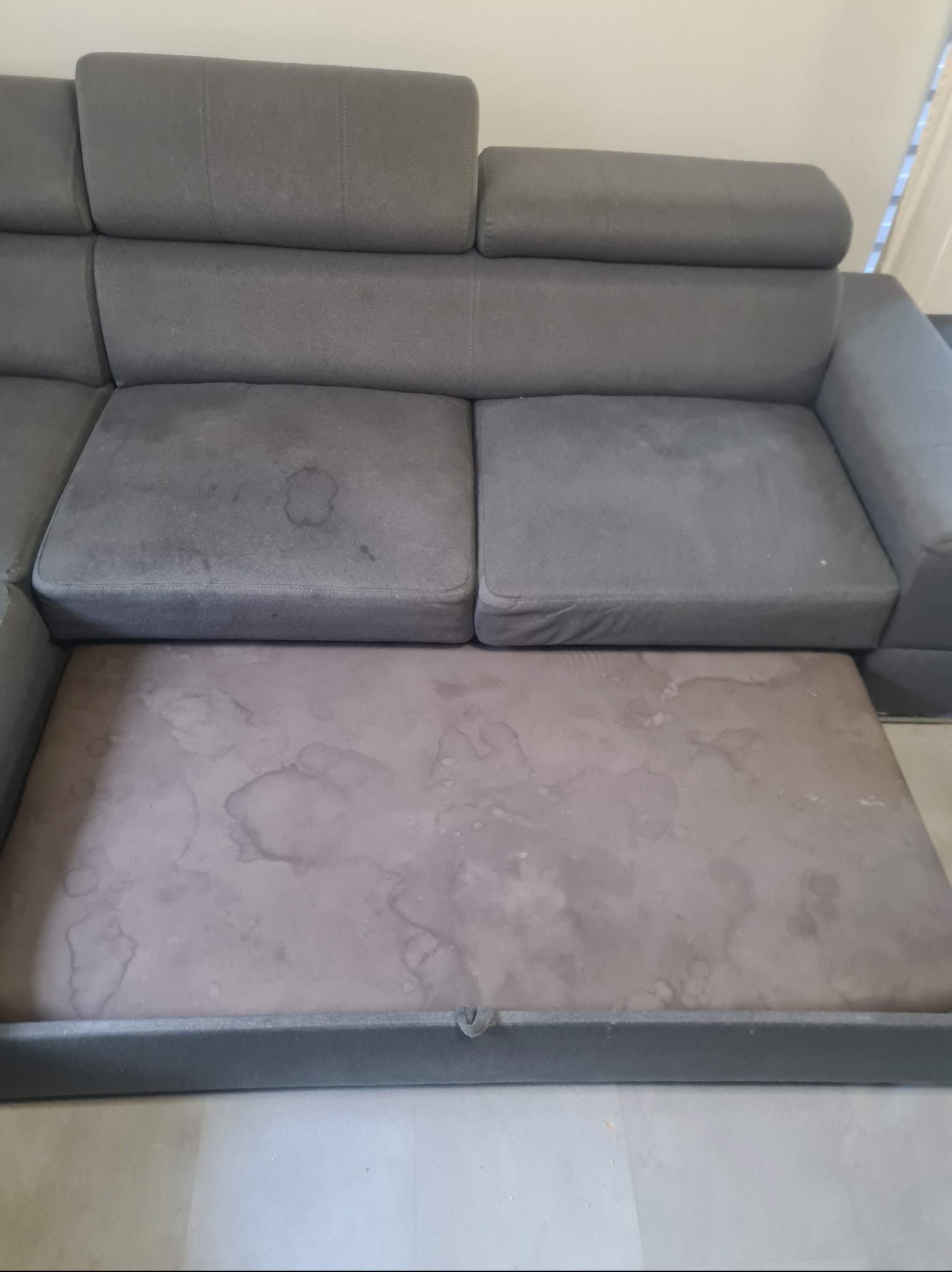 Szara kanapa z widocznymi plamami i rozłożonym siedziskiem.  Widoczne zabrudzenia na tapicerce. Potrzeba wyprania tapicerki.