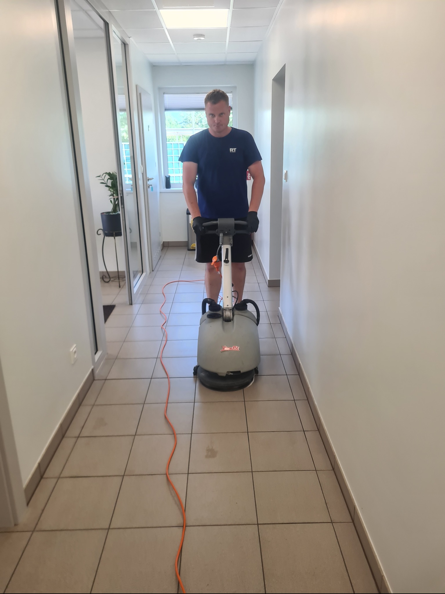 Pracownik z maszyną do czyszczenia podłóg w korytarzu biurowym. Widoczne płytki podłogowe i białe ściany. Pomarańczowy kabel maszyny. Ujęcie z przodu, na wprost.