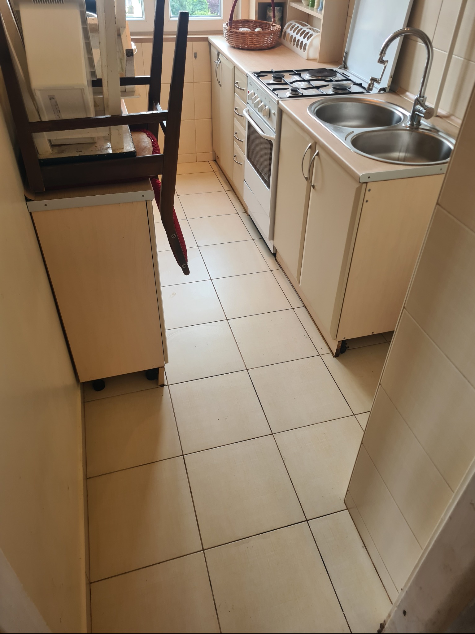 Kadr z kuchni z beżowymi płytkami podłogowymi, meblami i sprzętami. Na szafce stoi krzesło. Widoczny zlew, kuchenka i okno.