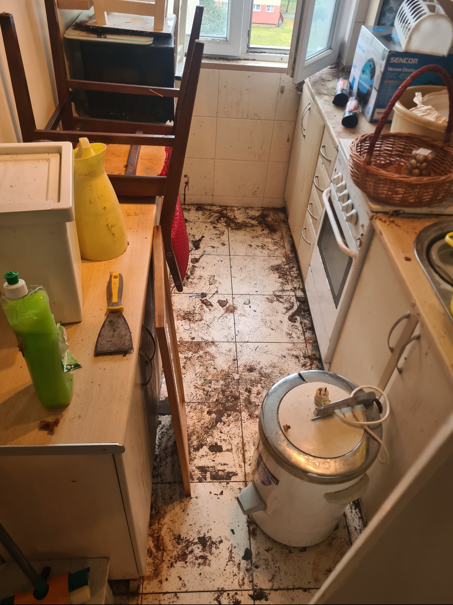 Zaniedbana kuchnia przed sprzątaniem: brudna podłoga, sprzęty i meble pokryte zaschniętym brudem. Widoczny sprzęt czyszczący i resztki po remoncie.
