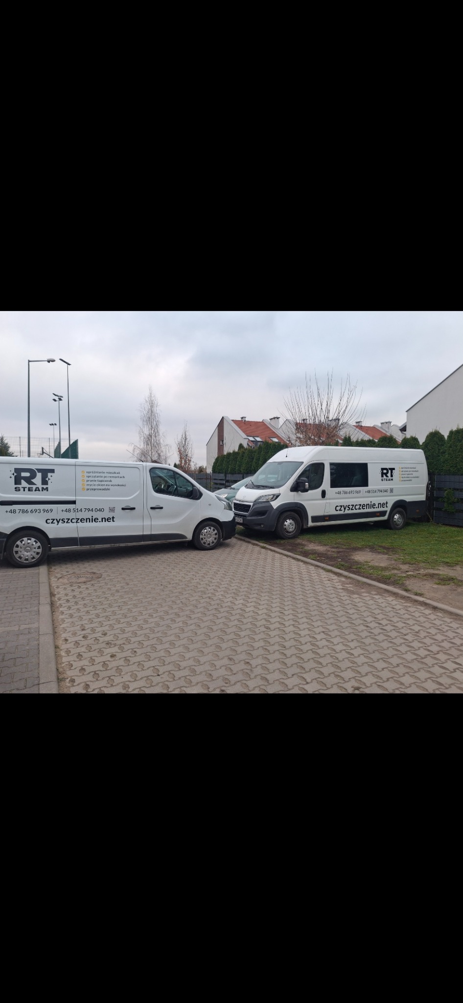 Dwa białe busy firmy czyszczącej RT Steam zaparkowane obok siebie na brukowanym podjeździe, z widocznymi numerami telefonów i adresem strony internetowej.