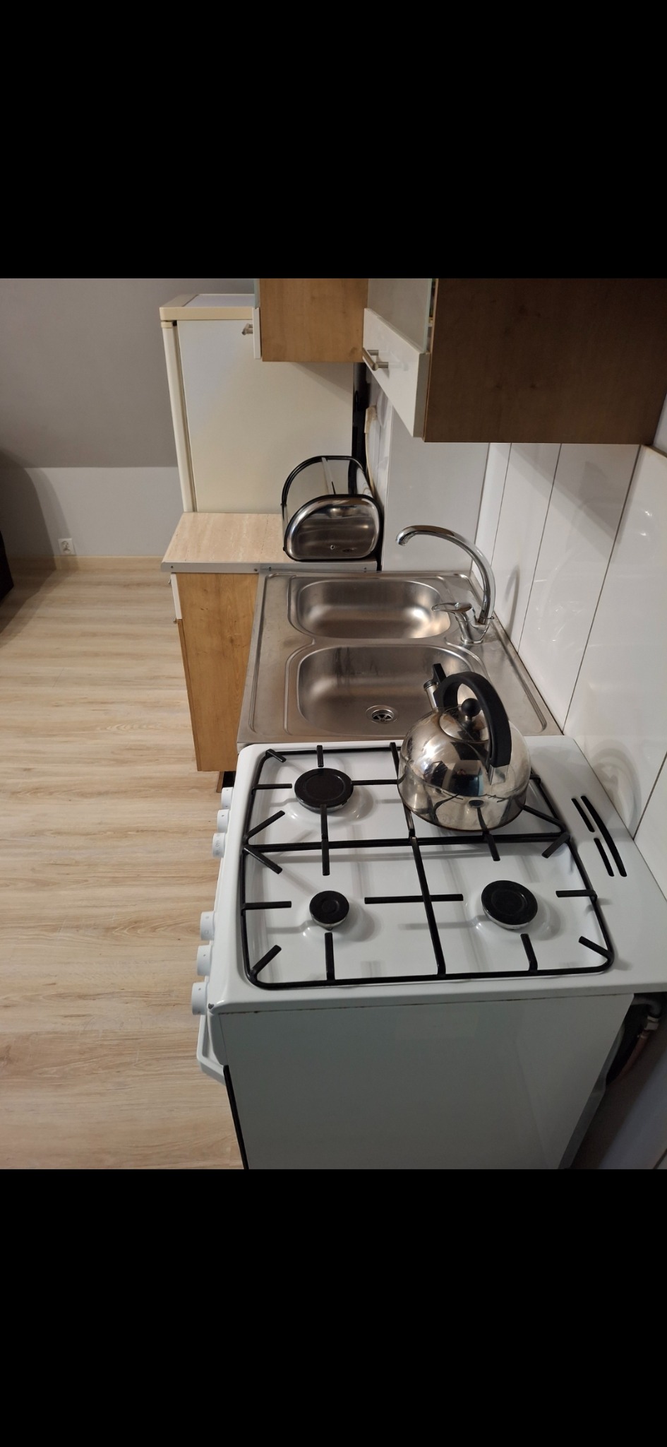 Umyta kuchnia w mieszkaniu na poddaszu: biała kuchenka z czajnikiem, dwukomorowy zlew, suszarka i lodówka. Jasne panele na podłodze, białe płytki na ścianie.