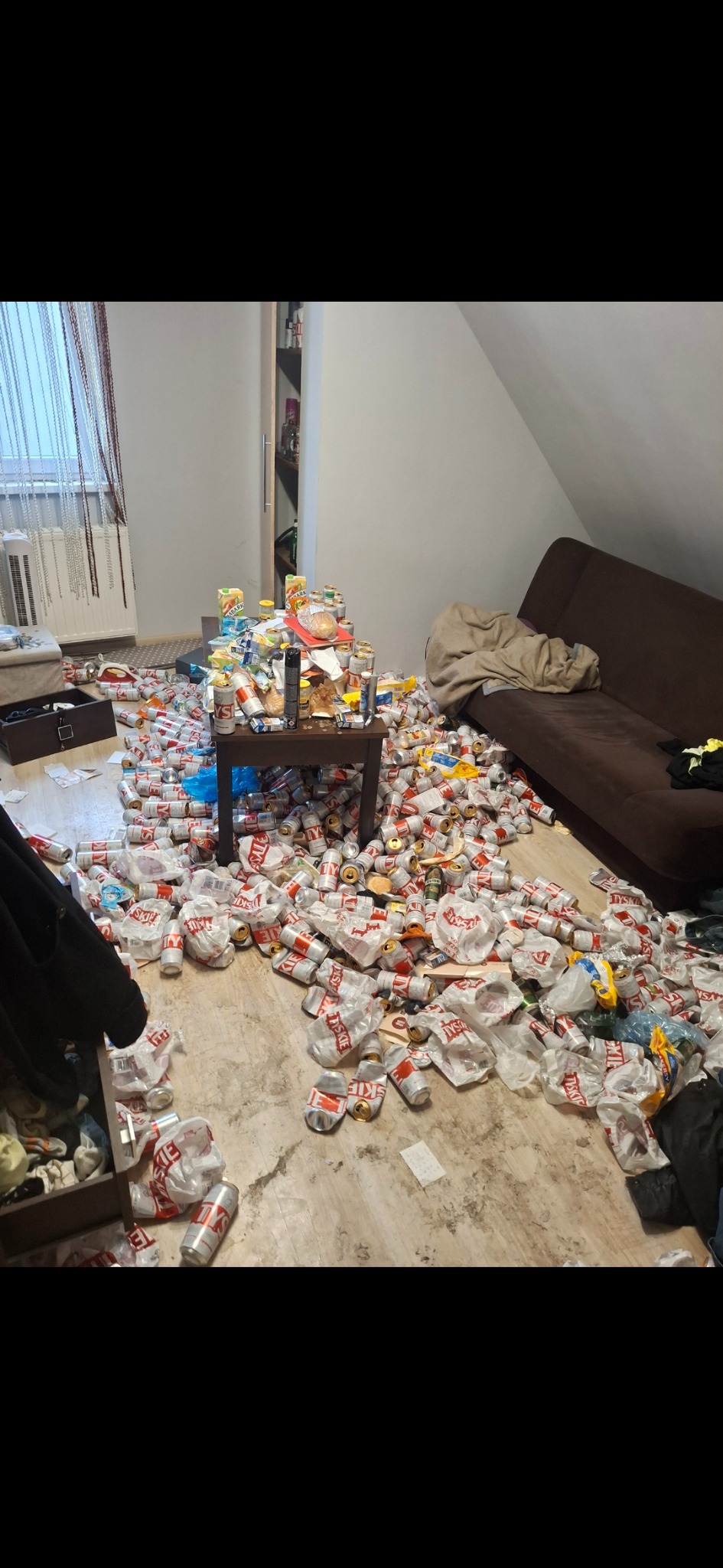 Zaniedbane poddasze z porozrzucanymi puszkami po piwie, śmieciami i brudną podłogą. Na stole resztki jedzenia i napojów. Widoczna sofa i okno.