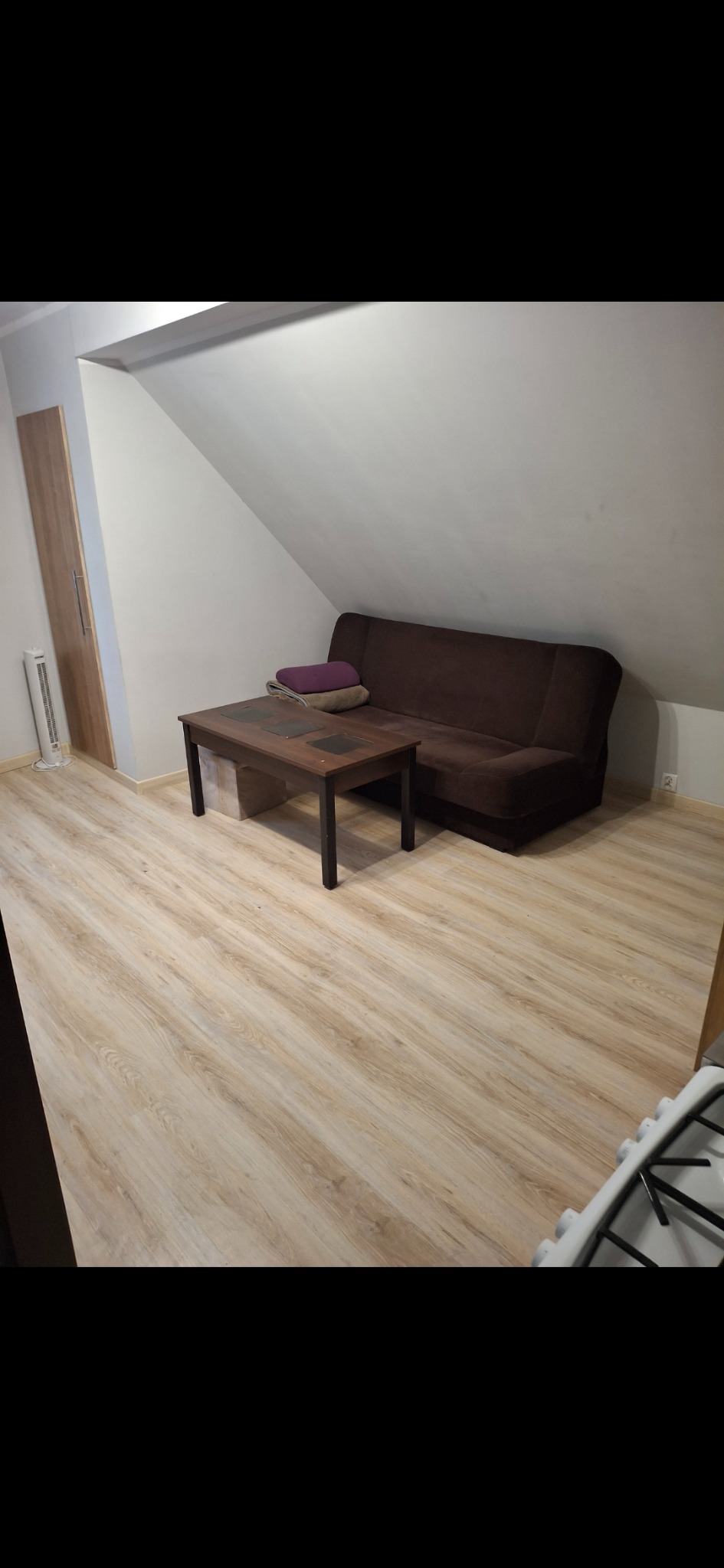 Uporządkowane poddasze ze skośnym sufitem, brązową sofą, drewnianym stolikiem i jasną podłogą. Widoczny fragment kuchenki.