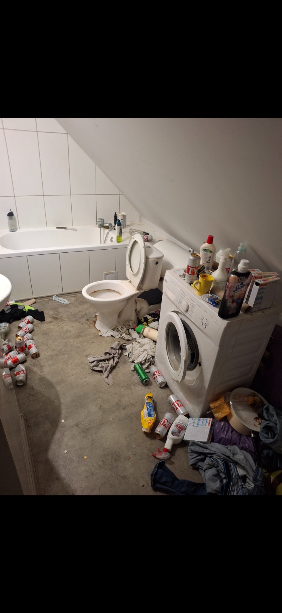 Zaniedbana łazienka na poddaszu z otwartą klapą toalety, stertą śmieci, puszek i ubrań obok pralki. Widok na wannę i umywalkę w tle.