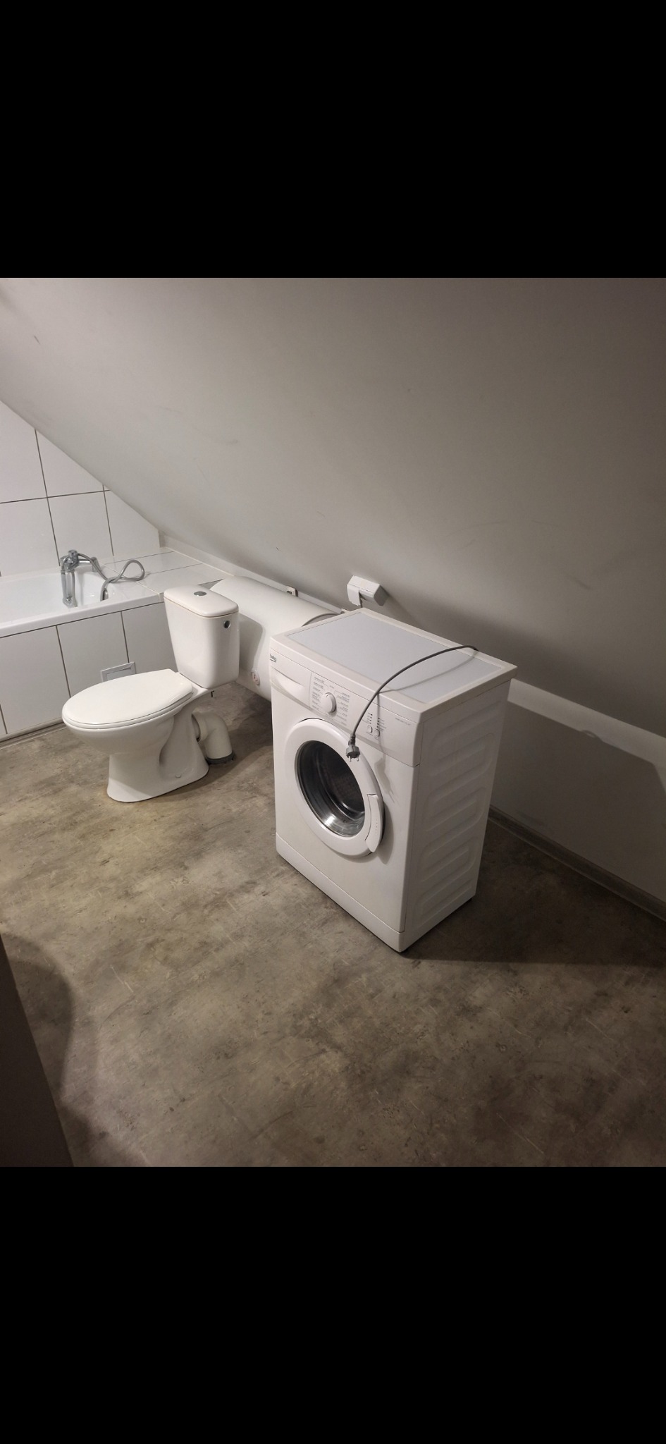 Ukończona łazienka na poddaszu ze skośnym sufitem, z białą wanną, toaletą i pralką. Podłoga betonowa. Widok na całe pomieszczenie.