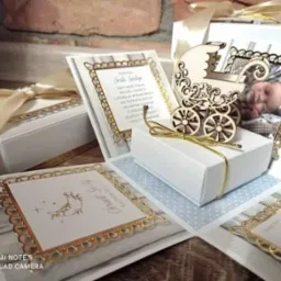 Otwarty exploding box z motywami chrztu, zawierający ozdobne kartki z życzeniami, drewniany wózek i zdjęcie śpiącego niemowlęcia, całość utrzymana w bieli i złocie.