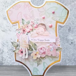 Ręcznie robiona kartka okolicznościowa w kształcie dziecięcego body, zdobiona motywami kwiatowymi, motylami, figurką śpiącego dziecka, królika i słonia, z napisem 'W dniu Twojego Święta'.