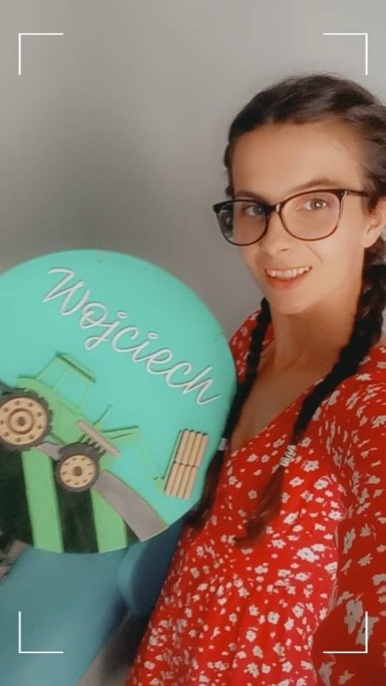 Uśmiechnięta kobieta w okularach trzyma okrągłą, turkusową dekorację z wyciętym traktorem i napisem 'Wojciech' wykonanym ozdobną czcionką.