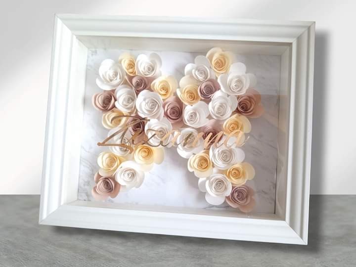 Oprawiona w białą ramę kompozycja z papierowych róż w odcieniach bieli, beżu i różu, ułożonych w kształt litery 'M' na marmurkowym tle, z napisem w kolorze złotym.