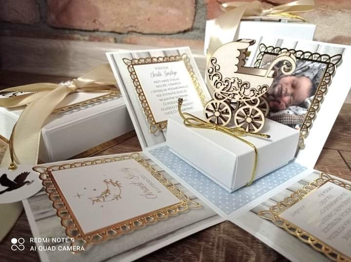 Otwarty exploding box z motywami chrztu, zawierający ozdobne kartki z życzeniami, drewniany wózek i zdjęcie śpiącego niemowlęcia, całość utrzymana w bieli i złocie.