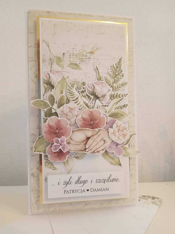 Ręcznie robiona kartka ślubna z motywem kwiatowym i dłońmi z pierścionkiem, umieszczona na tle z nutami i tekstem, z napisem '...i żyli długo i szczęśliwie. Patrycja & Damian'.