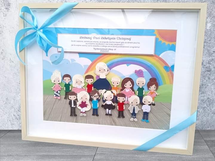 Ręcznie robiona pamiątka w ramce, przedstawiająca grupę dzieci i nauczycielkę w formie wyciętych postaci z papieru, z tęczą i słońcem w tle, dedykowana 'Kochanej Pani Katarzynie Szczęsnej'...