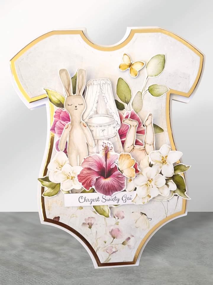 Ręcznie robiona kartka okolicznościowa z okazji Chrztu Świętego Gai, w kształcie dziecięcego body, zdobiona kwiatami, figurką królika i miniaturą łóżeczka, wykonana przez grafika z Nowej Wsi.