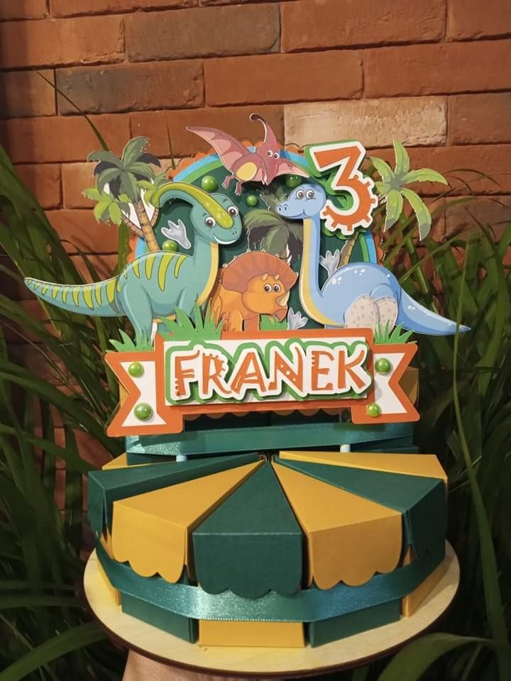 Trójpoziomowy tort urodzinowy z motywem dinozaurów i imieniem 'Franek' na ozdobnej tabliczce, na tle ceglanej ściany.