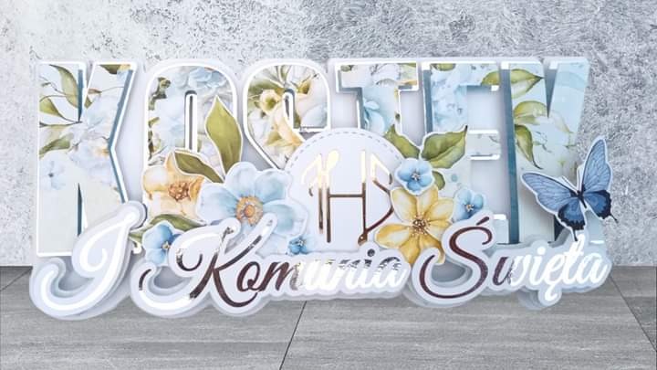 Dekoracja stołu na Pierwszą Komunię Świętą: przestrzenne litery z motywem kwiatowym i motylem, wykonane z papieru i kartonu.