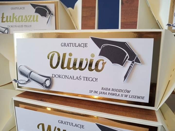 Ręcznie robione pudełko z kartką gratulacyjną dla Oliwii z okazji ukończenia szkoły, ozdobione złotymi napisami i motywami absolwenckimi, w tle widoczne inne podobne pudełka.