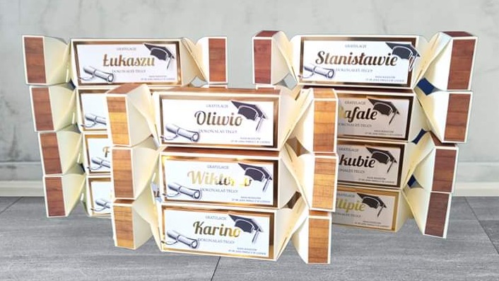 Personalizowane pudełka na upominki z imionami i motywem ukończenia szkoły, ułożone w piramidy, z widoczną fakturą drewna na bokach.