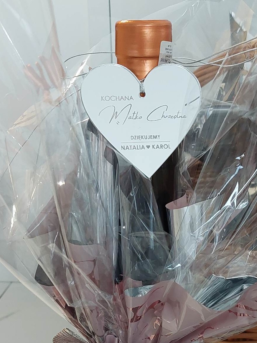 Prezent zapakowany w przezroczysty celofan z zawieszką w kształcie serca z napisem 'Kochana Matko Chrzestna. Dziękujemy. Natalia i Karol'. Widoczna butelka z miedzianą zakrętką.