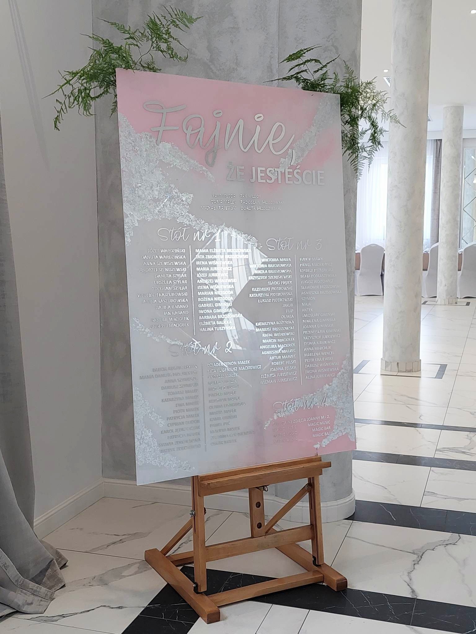 Elegancka tablica z listą gości weselnych, umieszczona na drewnianym sztalugie, z napisem 'Fajnie, że jesteście' i imionami gości w srebrnej czcionce na tle w odcieniach różu i srebra, udekorowana...