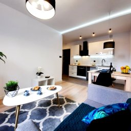 MARASKU Tomasz Żak - Jasne wnętrze mieszkania z otwartą kuchnią, widok na salon z sofą, stolikiem kawowym i dekoracjami roślinnymi, w tle biała zabudowa kuchenna z czarnym okapem i płytkami na ścianie.