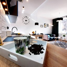 MARASKU Tomasz Żak - Nowoczesna kuchnia otwarta na salon, z białymi płytkami na ścianie, czarnym zlewem i płytą indukcyjną, na pierwszym planie taca z ziarnami kawy i kubkami w tropikalne liście, w tle stół jadalny...