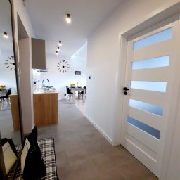 MARASKU Tomasz Żak - Nowoczesne wnętrze mieszkania z widokiem na kuchnię i jadalnię, szara płytka podłogowa, białe ściany, drzwi wewnętrzne z przeszkleniami i designerski zegar na ścianie.