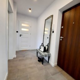 MARASKU Tomasz Żak - Jasny korytarz z beżowymi płytkami podłogowymi, białą ścianą, drewnianymi drzwiami wejściowymi, białymi drzwiami wewnętrznymi z przeszkleniami, wysokim lustrem opartym o ścianę, ławką w kratę...