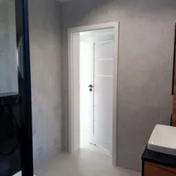 Nowoczesna łazienka z szarymi płytkami, białą umywalką na drewnianej szafce i otwartymi białymi drzwiami z czarną klamką.