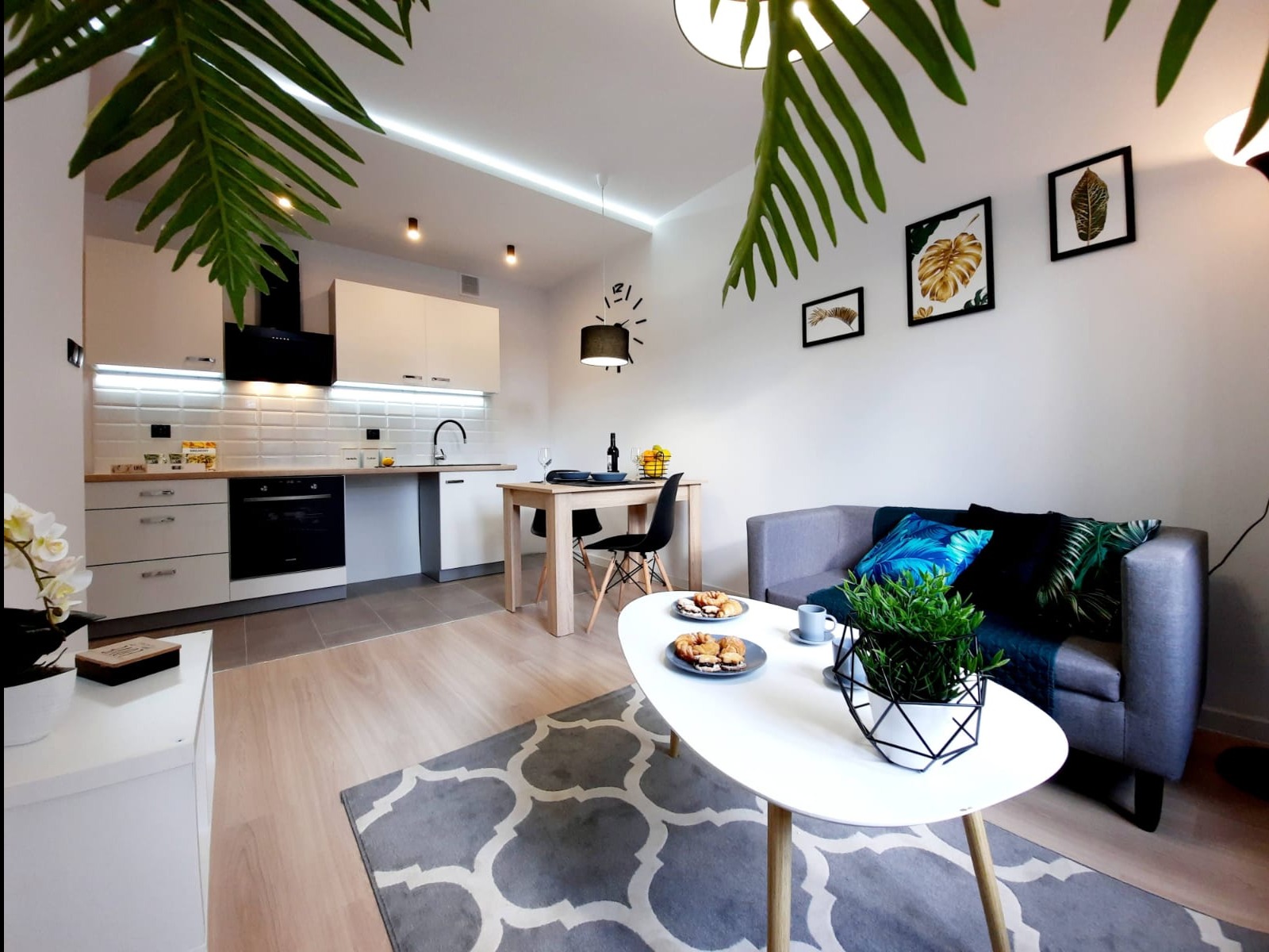 Nowoczesne wnętrze mieszkania z aneksem kuchennym, białymi meblami, drewnianym stołem, szarą sofą z poduszkami w tropikalne wzory i dekoracyjnymi roślinami w doniczkach