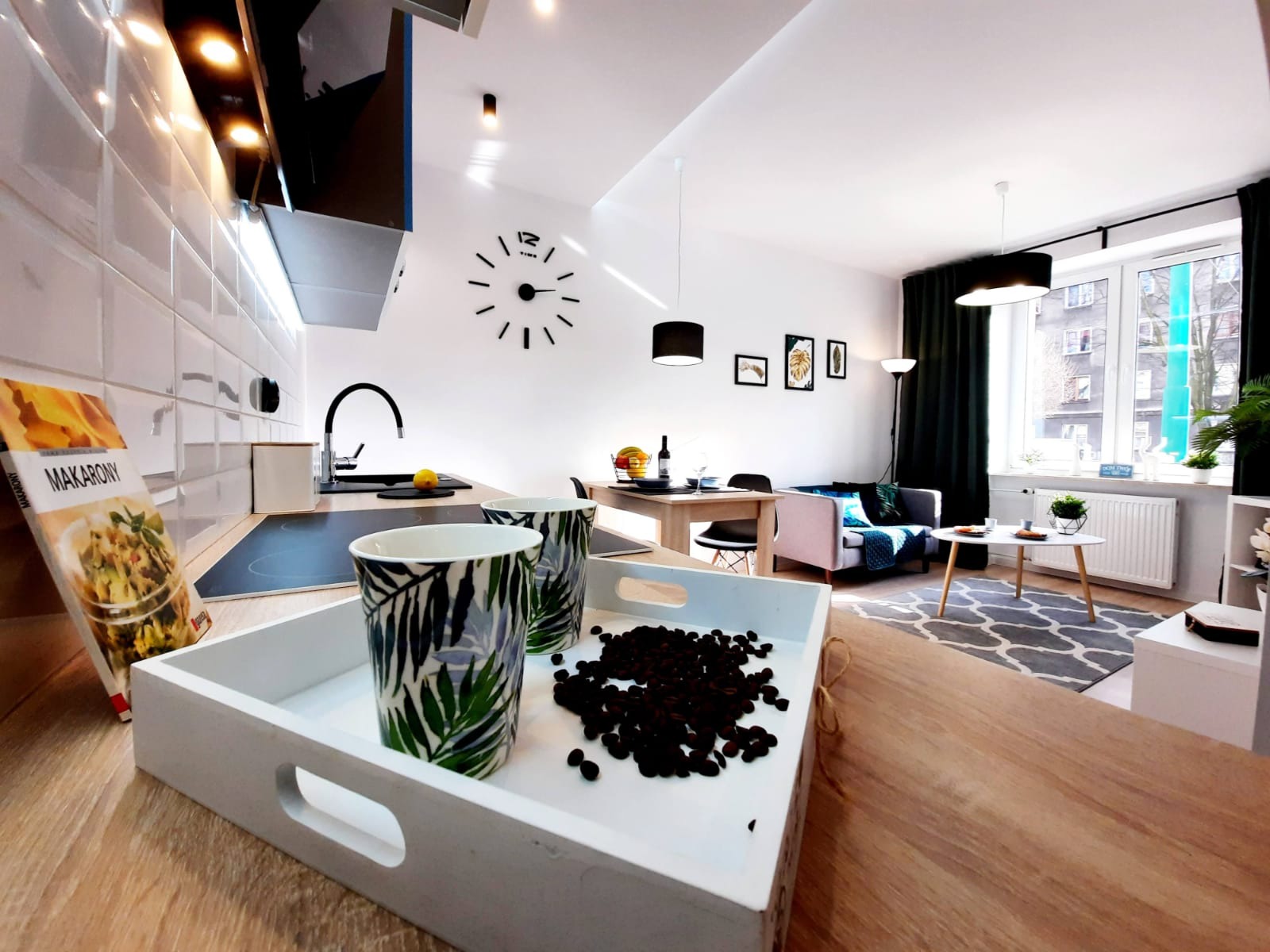 Nowoczesna kuchnia otwarta na salon, z białymi płytkami na ścianie, czarnym zlewem i płytą indukcyjną, na pierwszym planie taca z ziarnami kawy i kubkami w tropikalne liście, w tle stół jadalny...
