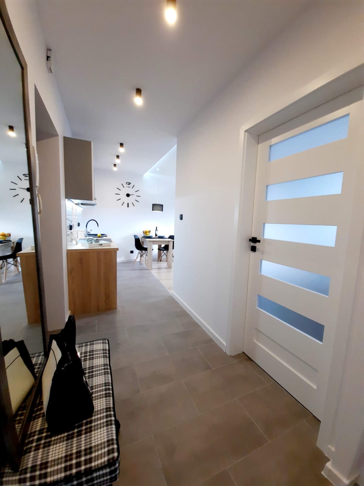 Nowoczesne wnętrze mieszkania z widokiem na kuchnię i jadalnię, szara płytka podłogowa, białe ściany, drzwi wewnętrzne z przeszkleniami i designerski zegar na ścianie.