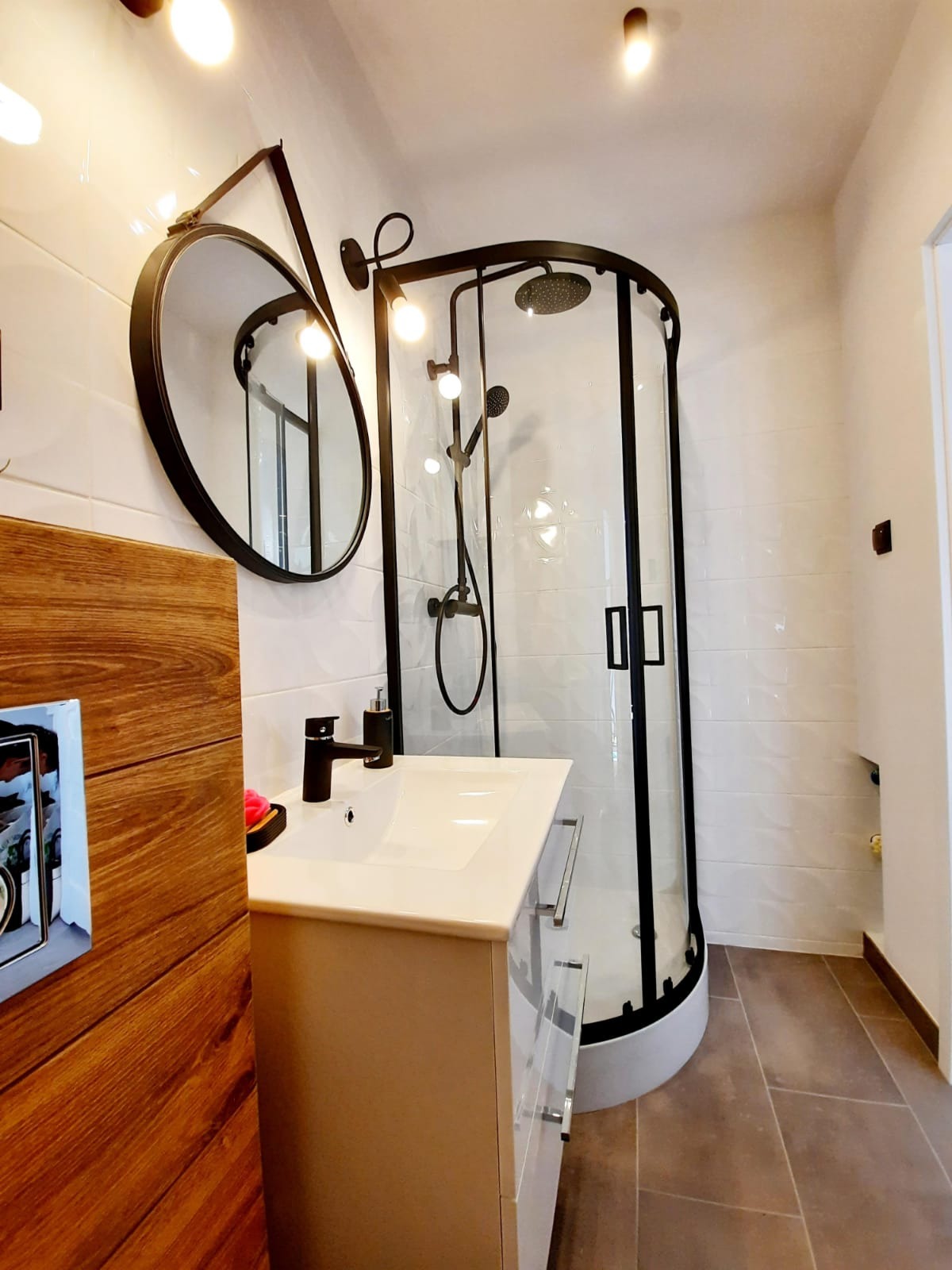 Nowoczesna łazienka z kabiną prysznicową w czarnej ramie, okrągłym lustrem na ścianie z białych płytek i umywalką z szafką w jasnym odcieniu, na podłodze brązowe płytki.