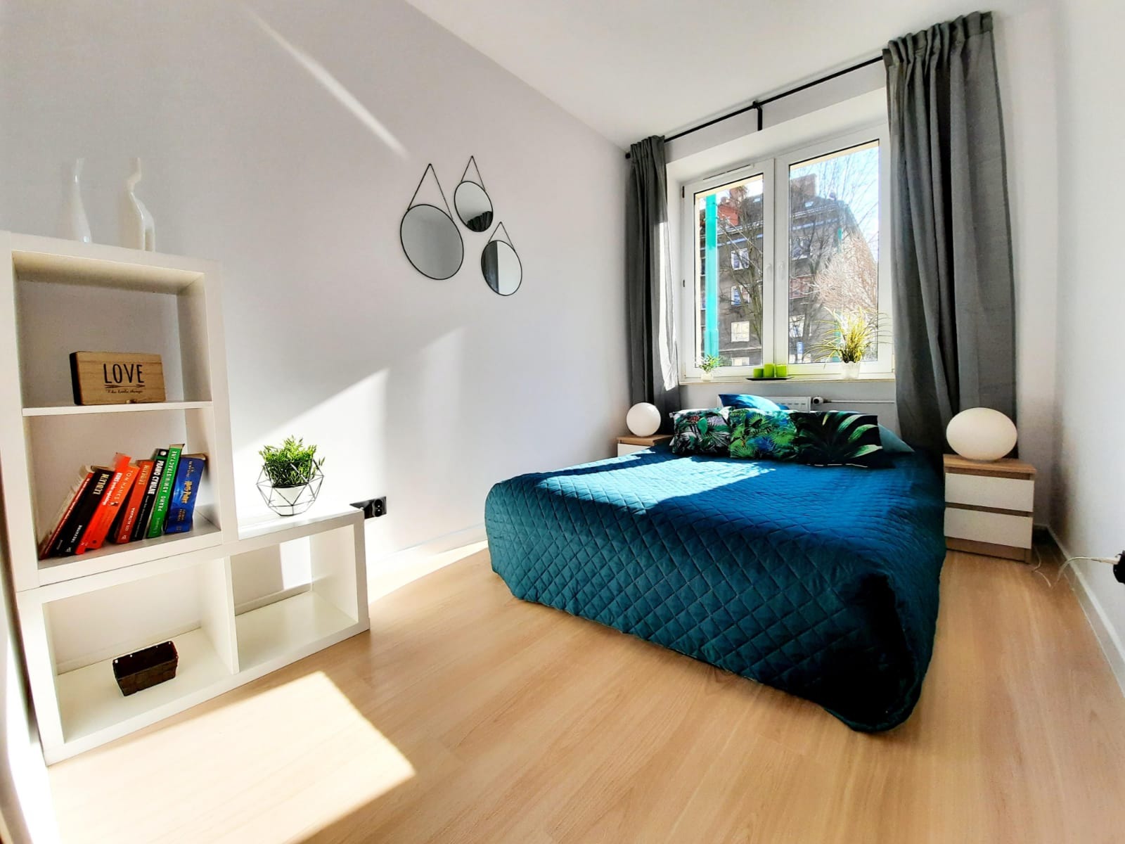 Jasna sypialnia z niebieską narzutą na łóżku, drewnianą podłogą, białym regałem z książkami i trzema okrągłymi lustrami na ścianie, oświetlona promieniami słonecznymi.
