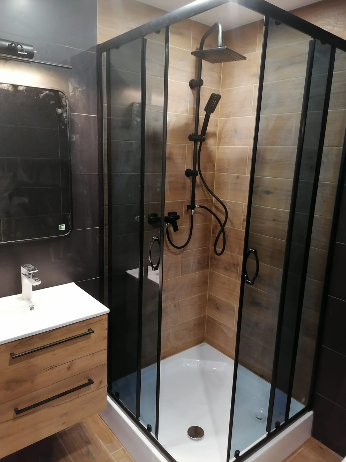 Nowoczesna łazienka z kabiną prysznicową typu walk-in z czarnymi profilami, drewnopodobne płytki na ścianach, umywalka nablatowa z szafką z drewnianymi frontami i czarnymi uchwytami.