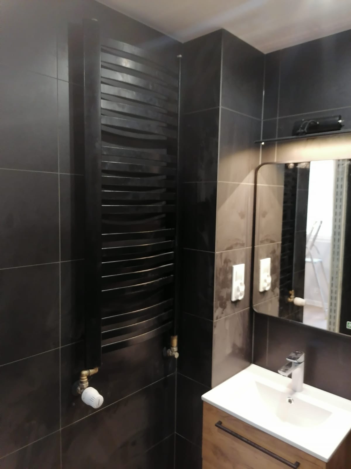 Ciemna łazienka z grzejnikiem drabinkowym o falistych szczeblach, umywalką nablatową i lustrem z widocznym fragmentem pomieszczenia w odbiciu.