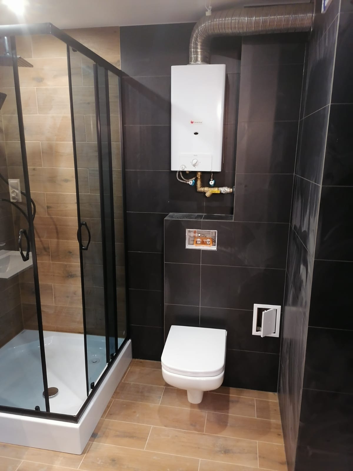 Nowoczesna łazienka z kabiną prysznicową z czarnymi ramami, podwieszaną toaletą, płytkami imitującymi drewno na podłodze i ciemnymi płytkami na ścianach, z zamontowanym na ścianie piecykiem gazowym.
