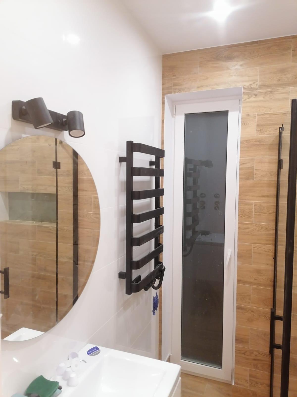 Nowoczesna łazienka po remoncie z owalnym lustrem, czarnym grzejnikiem drabinkowym, białymi płytkami na ścianie i imitacją drewna na części ściany oraz kabiną prysznicową.