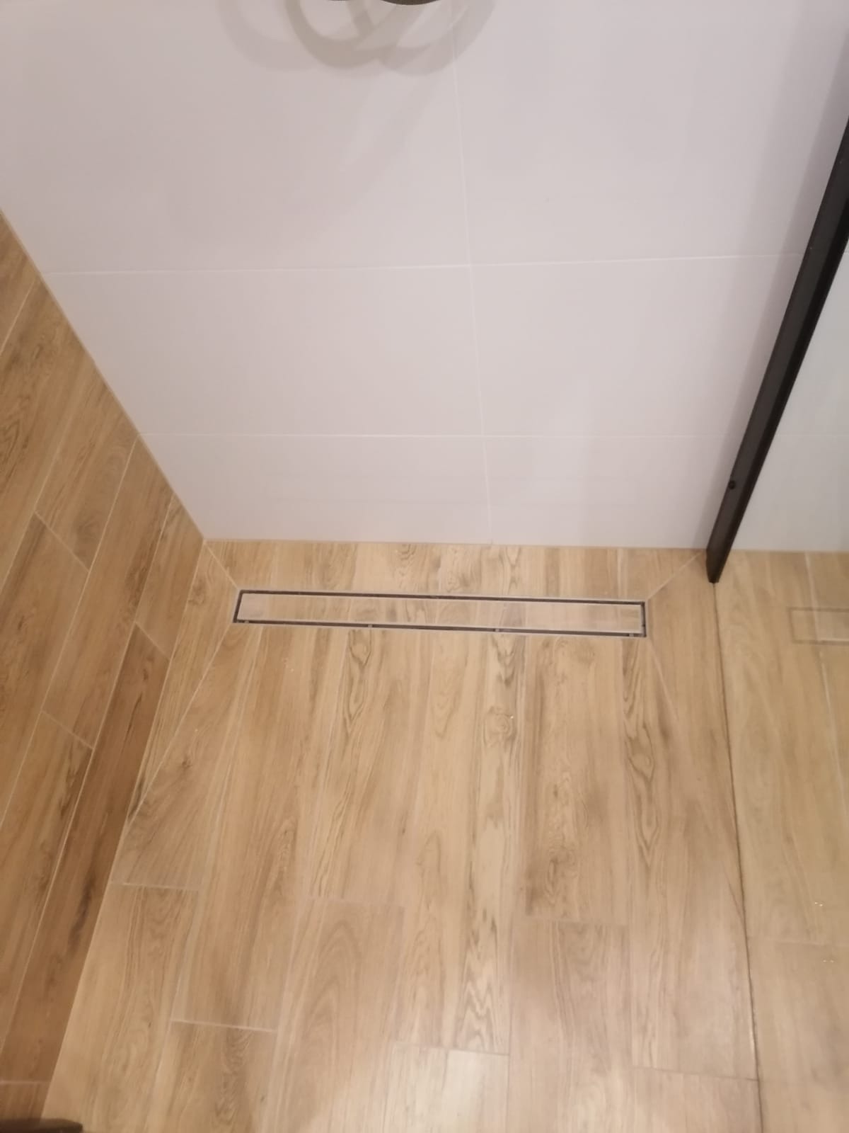 Wykończona kabina prysznicowa z odpływem liniowym, wyłożona płytkami imitującymi drewno, z białymi płytkami na ścianach i czarną ramą kabiny.