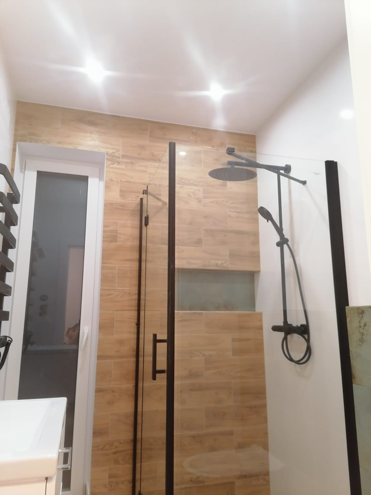 Nowoczesna kabina prysznicowa z deszczownicą i baterią w kolorze czarnym, zamontowana na ścianie wyłożonej płytkami imitującymi drewno, z widocznym oknem w białej ramie.