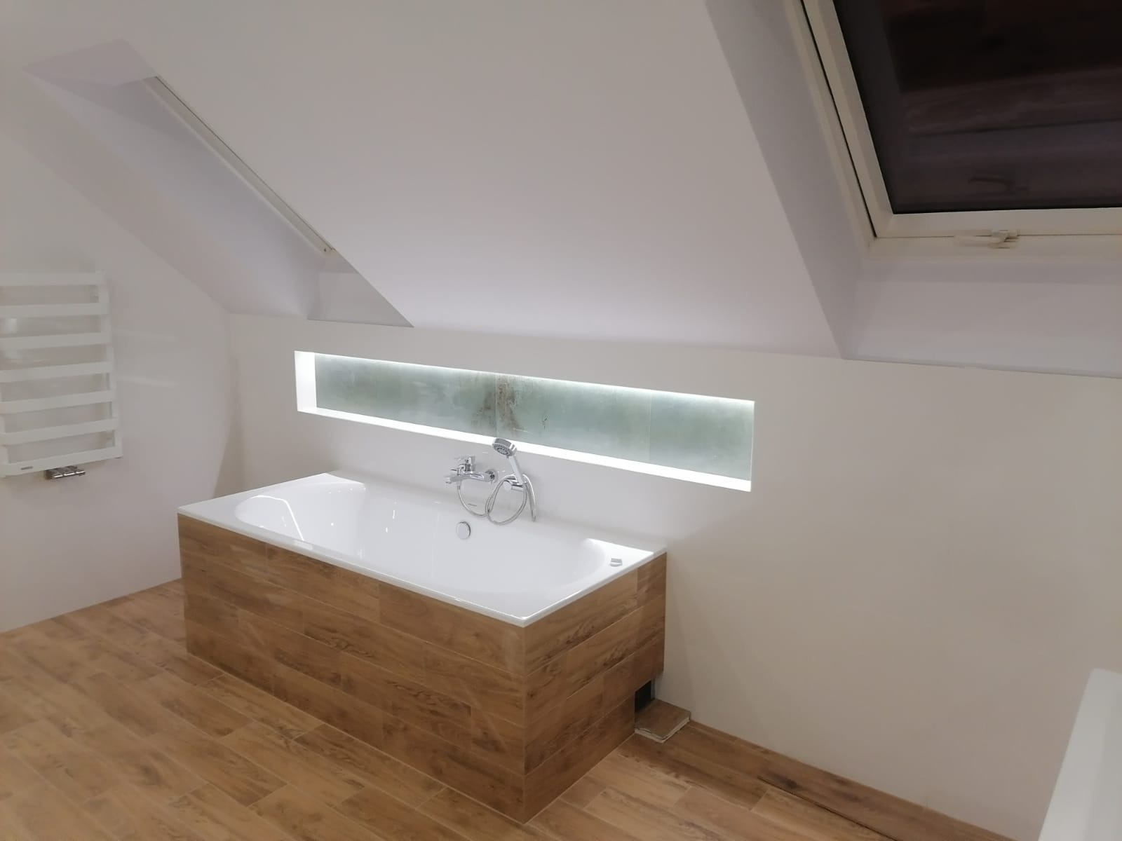 Biała wanna obudowana panelami imitującymi drewno, z chromowaną baterią i słuchawką prysznicową, umieszczona w łazience na poddaszu z oknem dachowym i podświetlaną wnęką nad wanną.