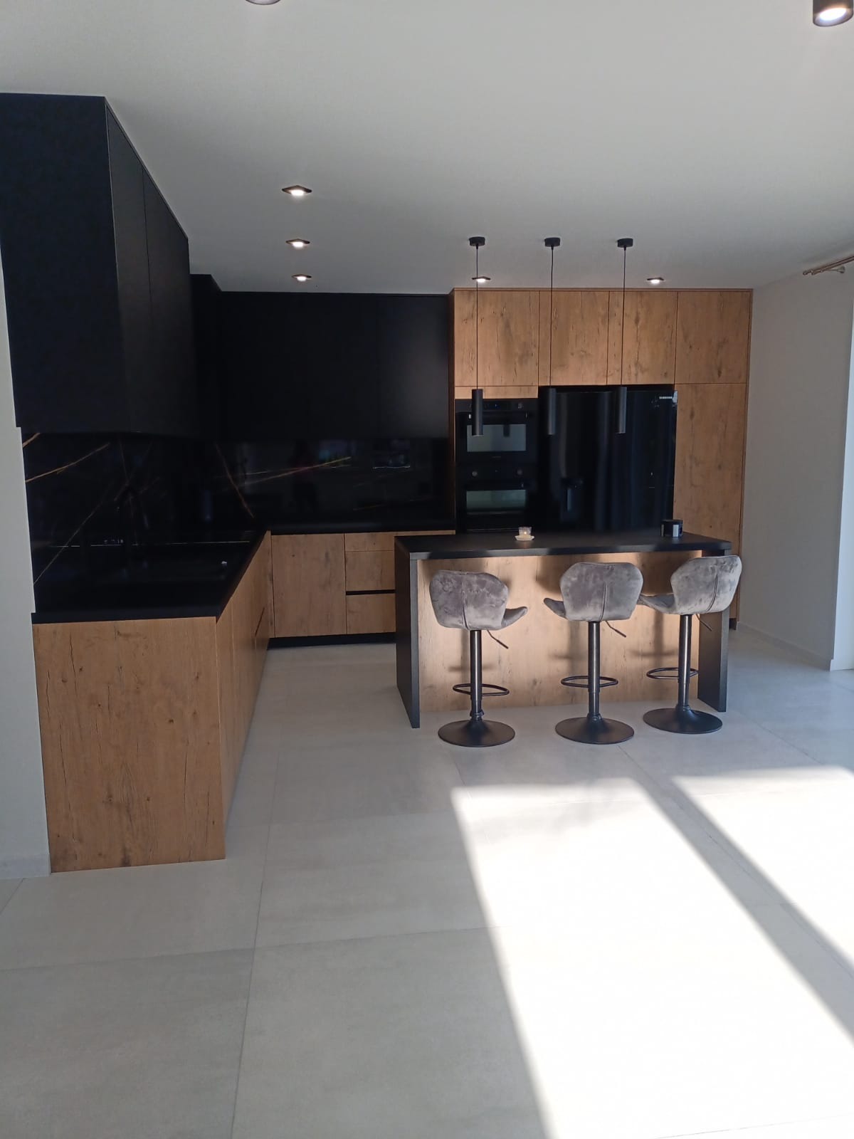 Nowoczesna kuchnia z drewnianymi frontami szafek, czarnym blatem i panelem ściennym imitującym marmur, wyspa z trzema szarymi hokerami na czarnych nogach.