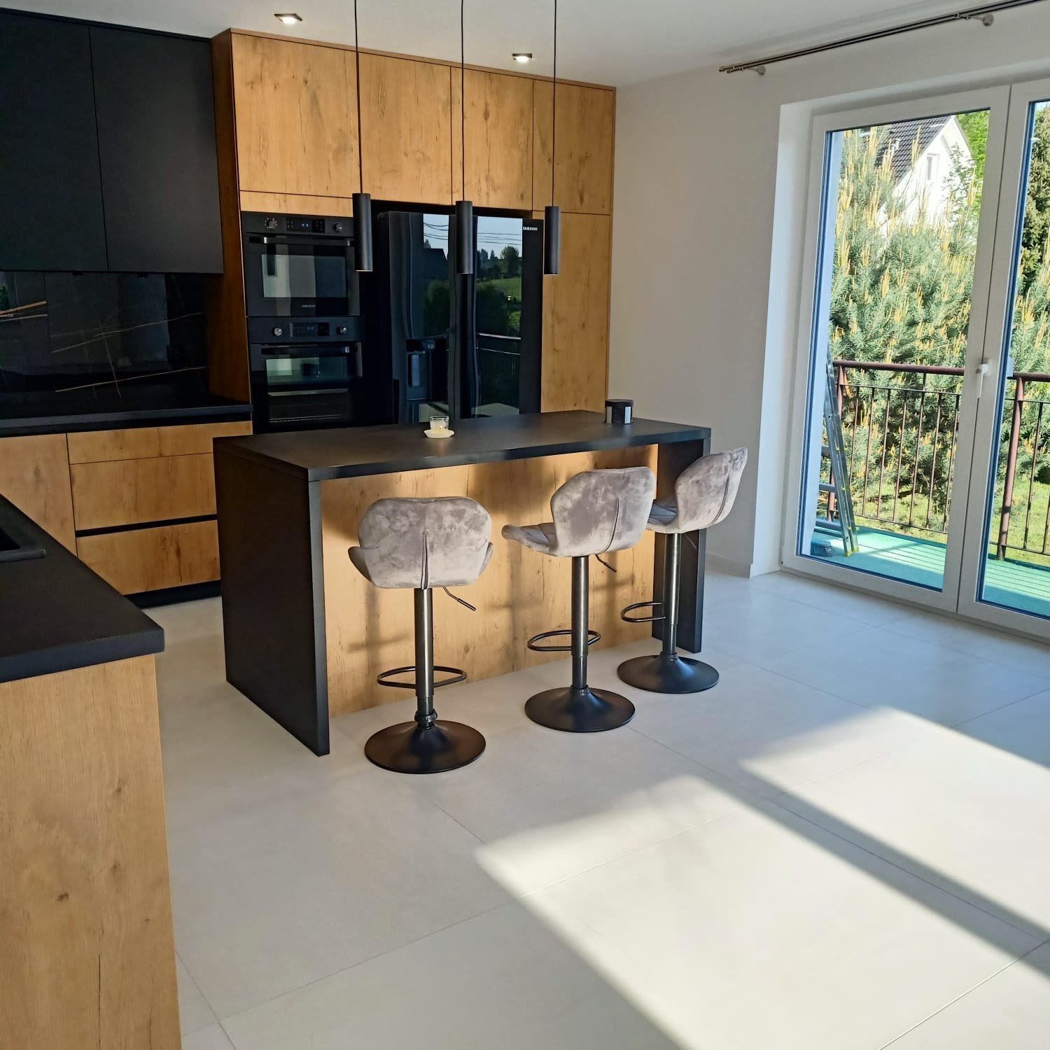 Nowoczesna kuchnia z drewnianymi frontami szafek, czarnymi elementami i wyspą z trzema szarymi hokerami na tle dużego okna balkonowego.