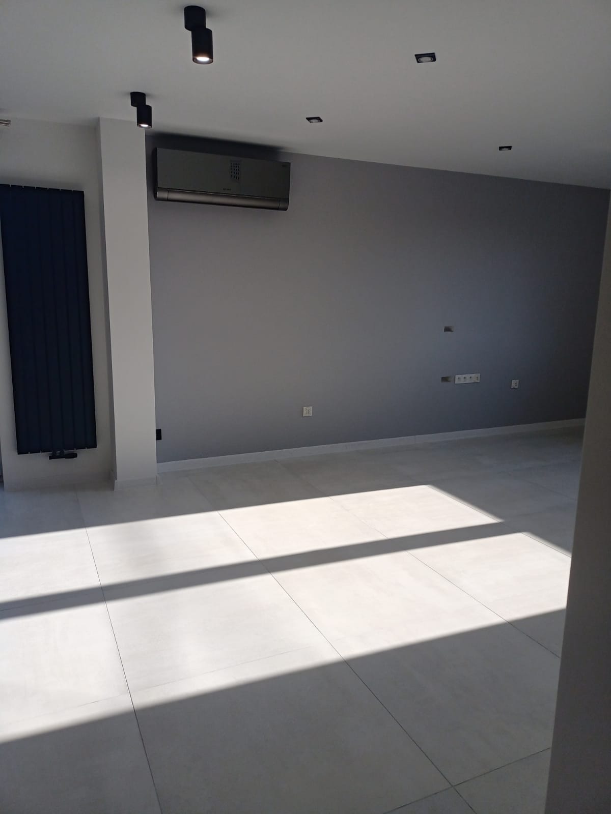 Pusty, szary pokój po remoncie z klimatyzacją, grzejnikiem panelowym i jasnymi płytkami podłogowymi, oświetlony promieniami słońca wpadającymi przez okno.