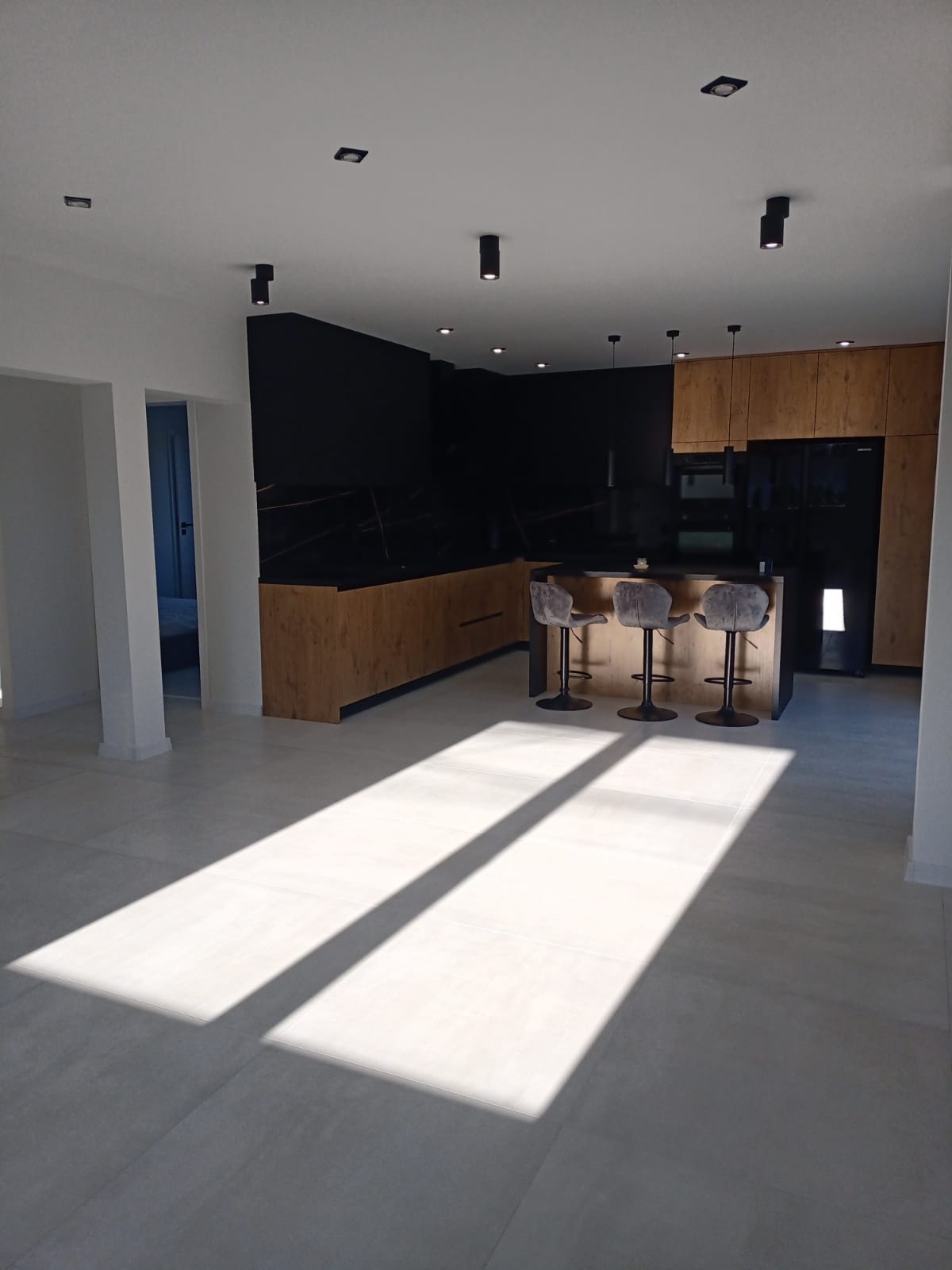 Nowoczesna kuchnia z wyspą, czarnymi frontami i drewnianymi akcentami, oświetlona promieniami słońca padającymi na jasną podłogę.